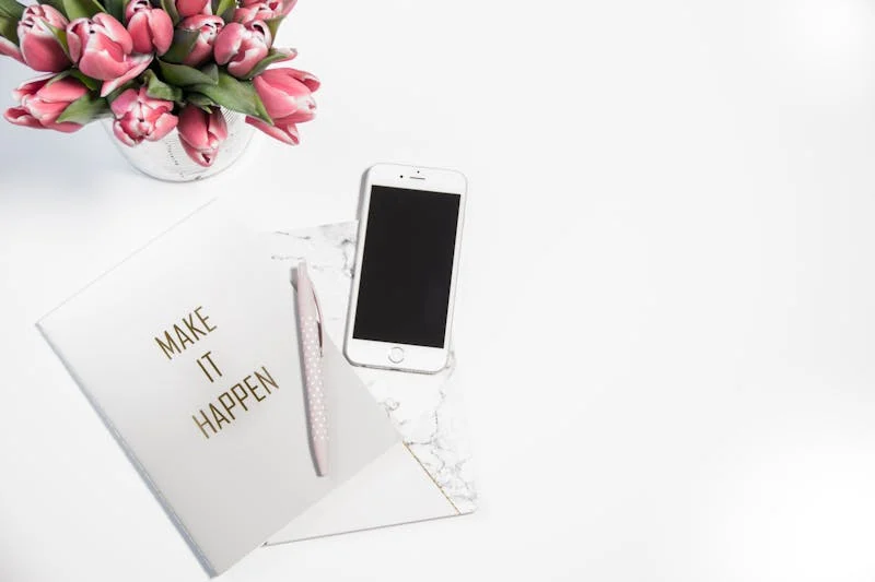 White background with pink tulips in the top left corner. A white cellphone that is off is in the center next to a white notebook that says make it happen on the cover in gold. a white pen is resting on top.