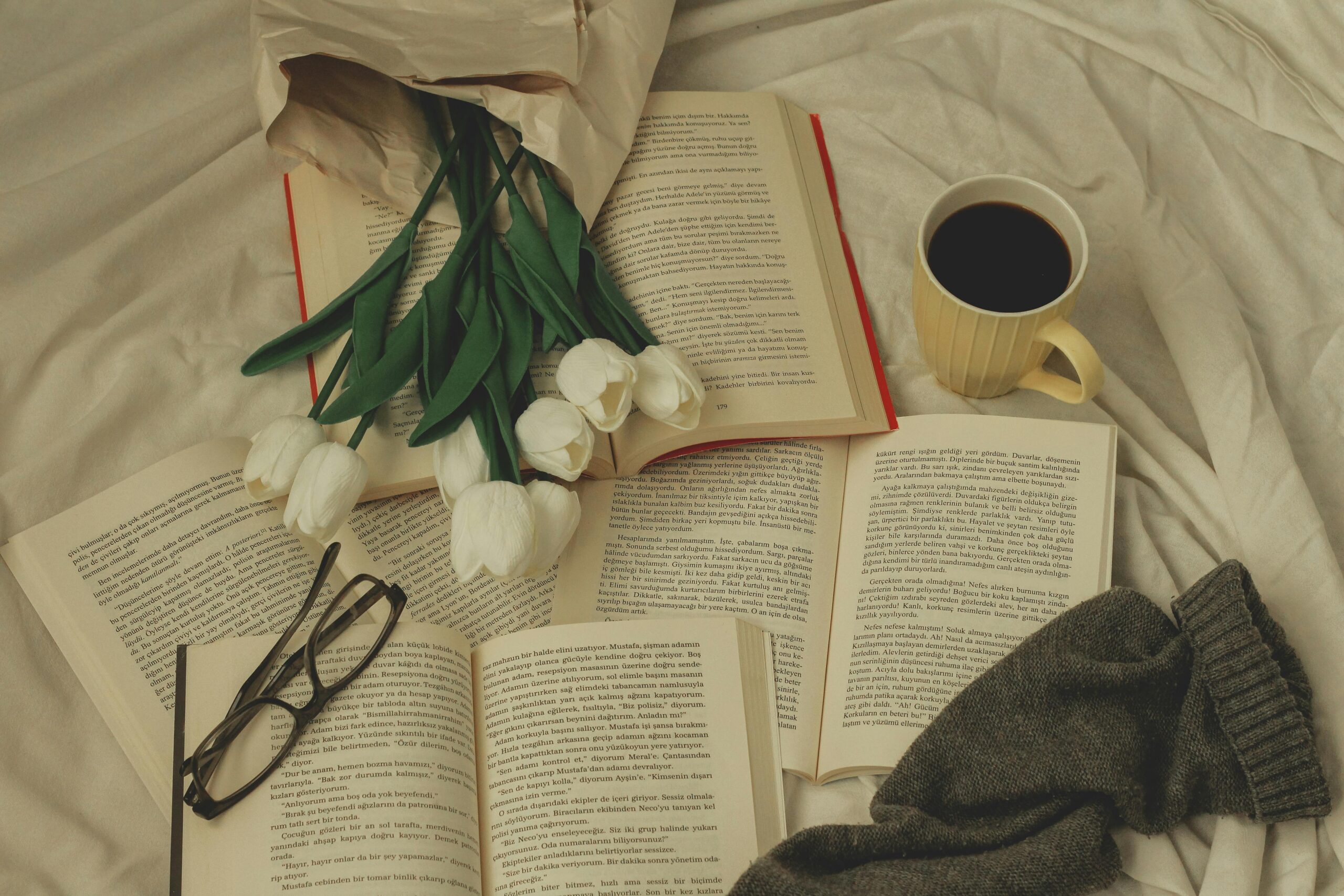 Flowers, white tulips, on top of open books on a bed with glasses and a yellow mug of coffee to the side. keywords used: bedroom cozy plants books mug.