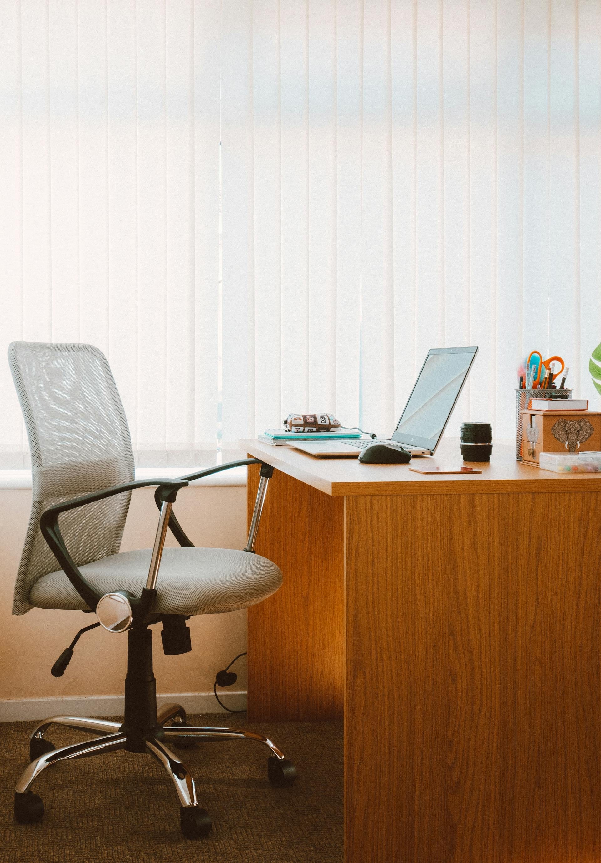 A white office chair is pulled out from an oak desk with a white laptop, writing utensils in a holder, and notebooks on the top in front of a window with white vertical blinds. Vibes are clean and organized.