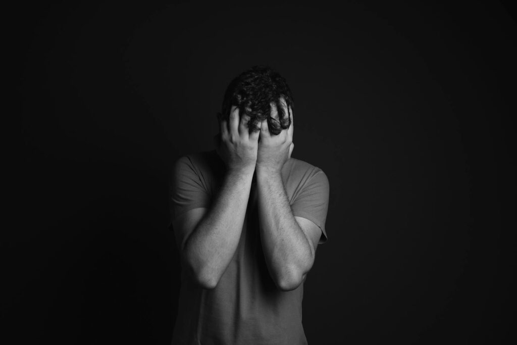 Black and white photo of a person burying their head into their hands out of grief. All black background. Vibes are gloomy and sad.